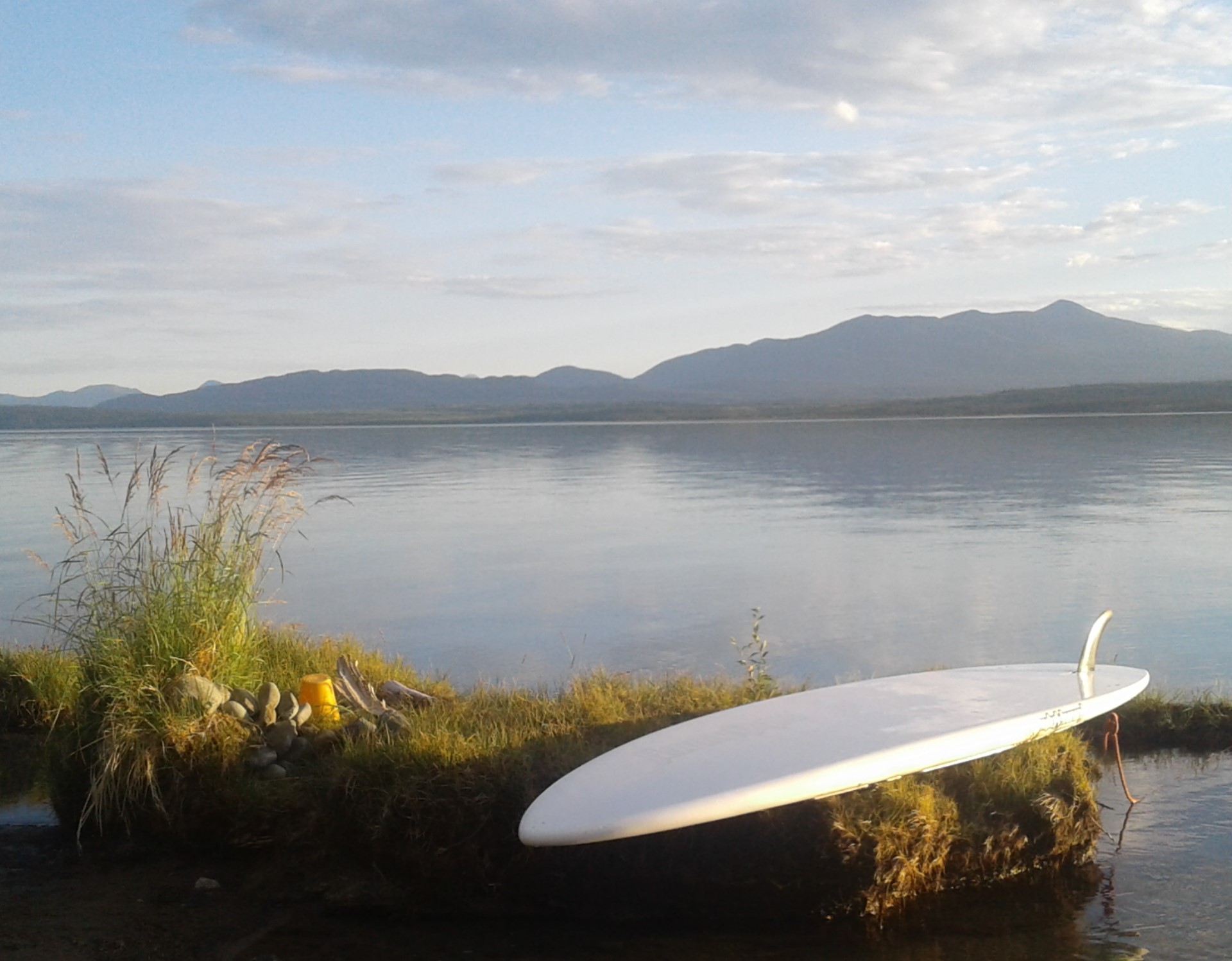 A white paddleboard sits on the shore of a lake with grey mountains in the background.